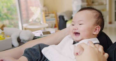 Bir bebek gülümsüyor ve bir insan tarafından tutuluyor. Bebek beyaz önlük giyiyor.