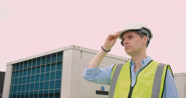 Beyaz kasketli ve yansıtıcı yelekli düşünceli bir mühendis endüstriyel ekipmanların yakınındaki bir inşaat sahasının mesafesine bakıyor.