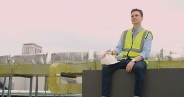 Sarı yelekli yakışıklı mühendis çatıdaki endüstriyel binanın pervazında oturuyor.