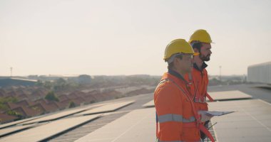 İki güvenlik ekipmanı mühendisi bir çatıdaki güneş panellerini inceliyor ve arka planda gün ışığında kentsel manzara var..