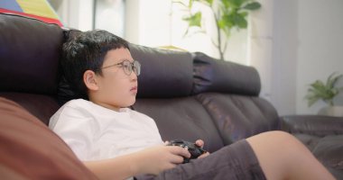 Asyalı çocuk oturma odasında kanepede oturuyor ve evde vakit geçirirken TV 'de oyun oynuyor.