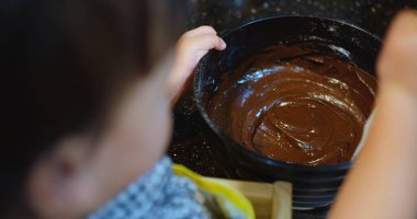 Bir çocuk anne babasıyla ev yapımı çikolatalı pasta yapıyor. Çocuk kasede bir kaşık krema tutuyor.