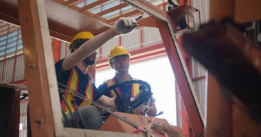 Sarı başlık ve güvenlik yelekleri giyen inşaat işçileri endüstriyel bir ortamda inşaat kaldırma mekanizması çalıştırmayı, mesleki güvenlik ve takım çalışmasına odaklanmayı tartışıyorlar.