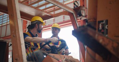 Sarı başlık ve güvenlik yelekleri giyen inşaat işçileri endüstriyel bir ortamda inşaat kaldırma mekanizması çalıştırmayı, mesleki güvenlik ve takım çalışmasına odaklanmayı tartışıyorlar.