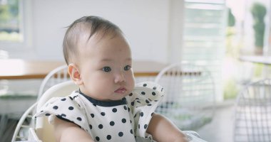 Bir bebek puantiyeli tişörtle yüksek bir sandalyede oturuyor. Bebek suratında bir somurtmayla kameraya bakıyor.