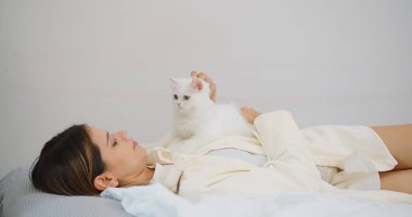 Çekici bir kadın bir yatakta uzanıyor, oturma odasında sevimli, tüylü, beyaz bir kediyle sevişiyor, ev sahibi evcil kedileri okşuyor, evcil hayvanlarla insanlar arasındaki ilişki, huzurlu ve rahatlatıcı.