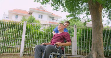 Bakıcı hemşire dışarıdaki bir evin bahçesinde tekerlekli sandalyede oturan Asyalı yaşlı bir adama yardım ediyor. Güneşli bir günün tadını çıkaran sakin bir açık havada şefkat ve destek gösteriyor.