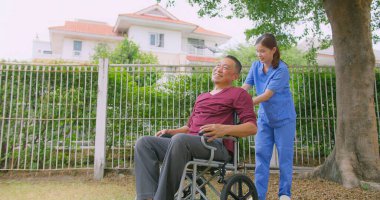Bakıcı hemşire dışarıdaki bir evin bahçesinde tekerlekli sandalyede oturan Asyalı yaşlı bir adama yardım ediyor. Güneşli bir günün tadını çıkaran sakin bir açık havada şefkat ve destek gösteriyor.