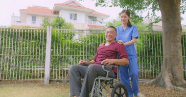 Bakıcı hemşire dışarıdaki bir evin bahçesinde tekerlekli sandalyede oturan Asyalı yaşlı bir adama yardım ediyor. Güneşli bir günün tadını çıkaran sakin bir açık havada şefkat ve destek gösteriyor.