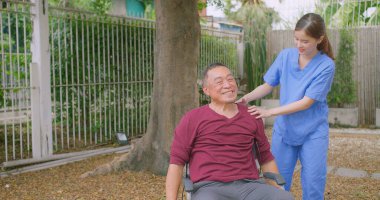 Bakıcı hemşire dışarıdaki bir evin bahçesinde tekerlekli sandalyede oturan Asyalı yaşlı bir adama yardım ediyor. Güneşli bir günün tadını çıkaran sakin bir açık havada şefkat ve destek gösteriyor.