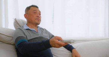 Yaşlı, yaşlı Asyalı bir adam, Beyaz Perdeli Parlak Oturma Odası 'nda elinde uzaktan kumandayla koltukta rahatça oturuyor.