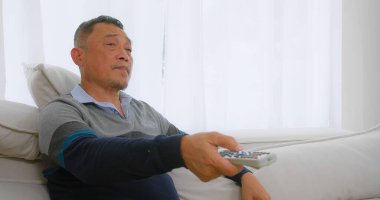 Yaşlı, yaşlı Asyalı bir adam, Beyaz Perdeli Parlak Oturma Odası 'nda elinde uzaktan kumandayla koltukta rahatça oturuyor.