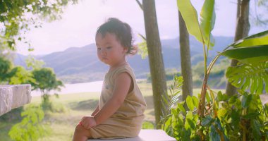 Güneşli bir günde, açık bir arka planda canlı yeşillikler ve uzak dağlarla çevrili doğada oturan Asyalı bir çocuğun huzurlu bir sahnesi.