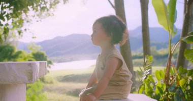 Güneşli bir günde, açık bir arka planda canlı yeşillikler ve uzak dağlarla çevrili doğada oturan Asyalı bir çocuğun huzurlu bir sahnesi.