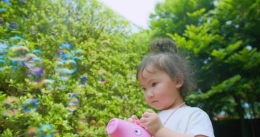 Mucize ifadesine sahip genç bir kız, arka planda yüzen kabarcıklar ve yeşilliklerle çevrili bir balon üfleyicisiyle açık havada oynuyor, çocukluğun masumiyetini ve neşesini doğal ışıkta yakalıyor..