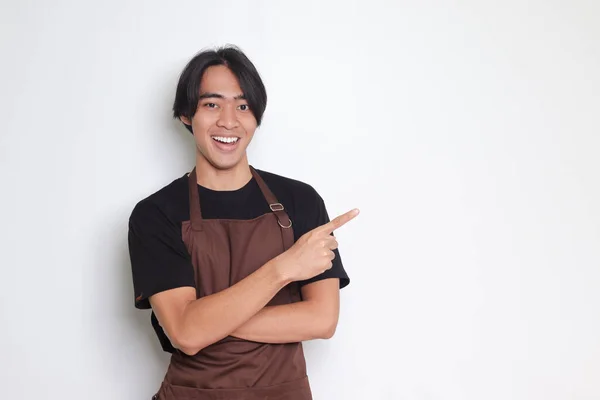 stock image Portrait of attractive Asian barista man in brown apron showing product, pointing at something with hands. Advertising concept. Isolated image on white background