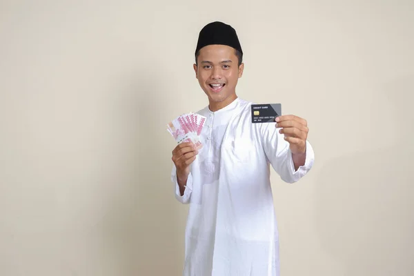 亚洲有魅力的穆斯林男子 身穿白衬衫 头戴一顶小帽 头戴十万卢比的肖像 还带着信用卡 灰色背景上的孤立图像 — 图库照片