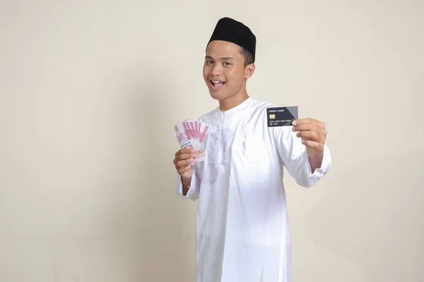 亚洲有魅力的穆斯林男子 身穿白衬衫 头戴一顶小帽 头戴十万卢比的肖像 还带着信用卡 灰色背景上的孤立图像 — 图库照片