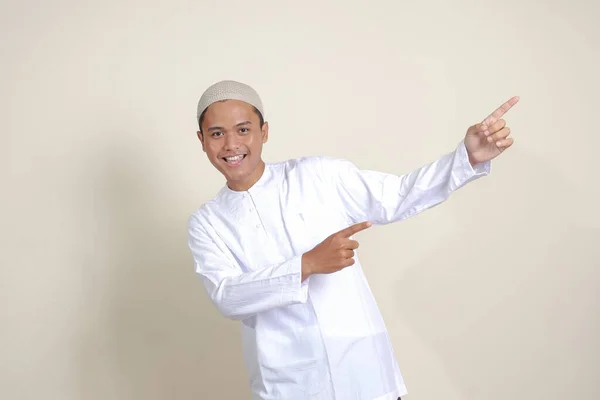 亚洲有魅力的穆斯林男子的画像 身穿白衬衫 头戴小帽 展示产品 手指头指向侧面 广告概念 灰色背景上的孤立图像 — 图库照片