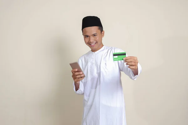 身穿白衬衫 头戴便帽 手持手机并出示信用卡的有魅力的亚洲穆斯林男子的画像 灰色背景上的孤立图像 — 图库照片