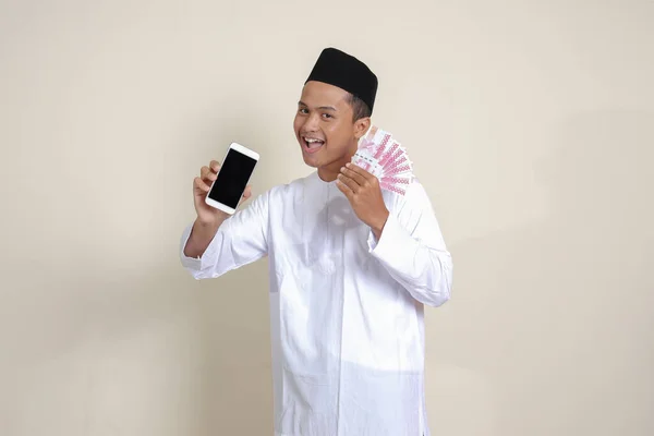 亚洲有魅力的穆斯林男子的画像 身穿白衬衫 头戴便帽 展示了十万卢比 同时展示了空白屏幕手机 金融和购物概念 灰度上的孤立图像 — 图库照片