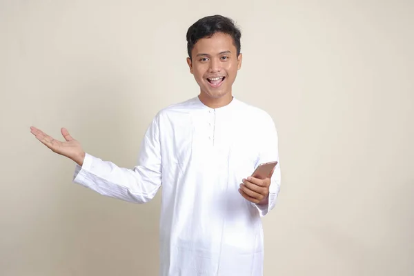 亚洲有魅力的穆斯林男子身穿白衬衫 手持手机 脸上挂着微笑的表情 同时将手指指向旁边的画像 广告概念 灰色背景上的孤立图像 — 图库照片