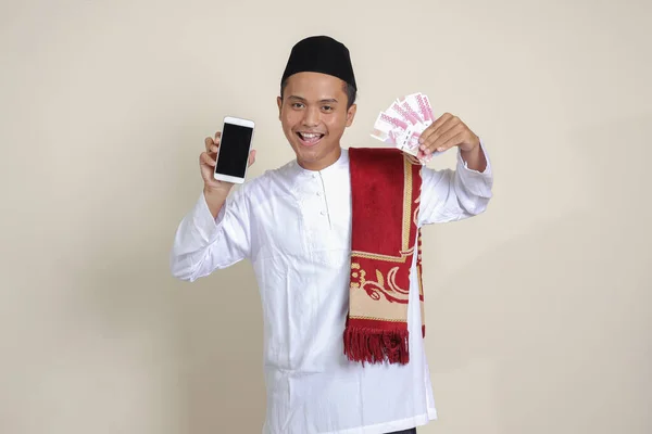 亚洲有魅力的穆斯林男子的画像 身穿白衬衫 头戴便帽 展示了十万卢比 同时展示了空白屏幕手机 金融和购物概念 灰度上的孤立图像 — 图库照片
