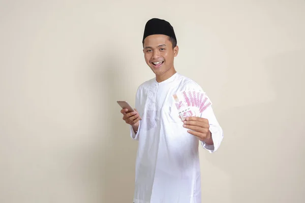 一个穿着白衬衫的有魅力的亚洲穆斯林男子的画像 在使用手机时显示出10万卢比 金融和储蓄概念 灰色背景上的孤立图像 — 图库照片