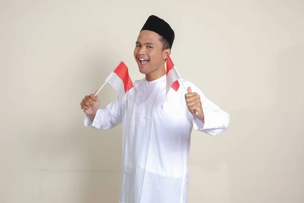 亚洲有魅力的穆斯林男子 身穿白衬衫 头戴便帽 举着印度尼西亚国旗 同时举起拳头 庆祝胜利 灰色背景上的孤立图像 — 图库照片