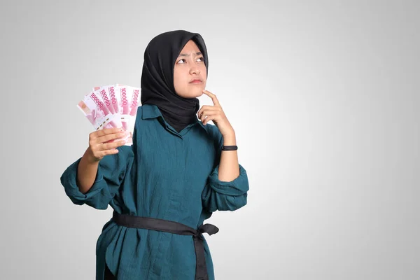 Portrait of confused Asian hijab woman in casual outfit showing one hundred thousand rupiah while thinking with hand on chin. Financial and savings concept. Isolated image on white background
