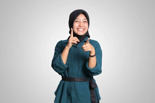 Portrait of excited Asian hijab woman in casual outfit standing against white background, showing product and pointing with hand and finger to the side. Advertising concept.