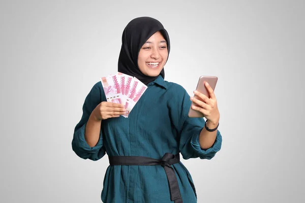 Portrait of excited Asian hijab woman in casual outfit showing one hundred thousand rupiah while holding a mobile phone. Financial and savings concept. Isolated image on white background