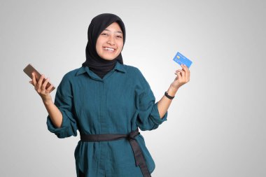 Tesettürlü neşeli Asyalı Müslüman kadın portresi, cep telefonu tutarken kredi kartını gösteriyor. Finansal ve tasarruf konsepti. Beyaz arkaplanda izole edilmiş resim