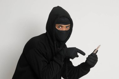 Siyah kapüşonlu ve maskeli gizemli bir adamın portresi cep telefonunda hackleme faaliyetleri yapıyor, elinde akıllı telefon tutan bir bilgisayar korsanı. Gri arkaplanda izole edilmiş resim