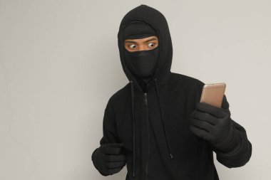 Siyah kapüşonlu ve maskeli gizemli bir adamın portresi cep telefonunda hackleme faaliyetleri yapıyor, elinde akıllı telefon tutan bir bilgisayar korsanı. Gri arkaplanda izole edilmiş resim