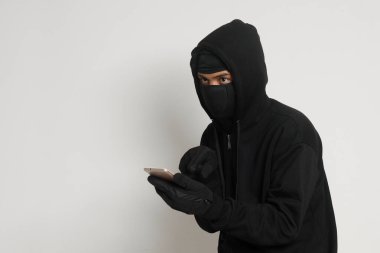 Siyah kapüşonlu ve maskeli gizemli bir adamın portresi cep telefonunda hackleme faaliyetleri yapıyor, elinde akıllı telefon tutan bir bilgisayar korsanı. Gri arkaplanda izole edilmiş resim