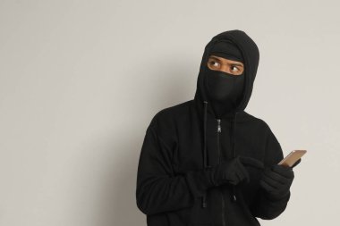 Siyah kapüşonlu ve maskeli gizemli bir adamın portresi cep telefonunda hackleme faaliyetleri yapıyor, elinde akıllı telefon tutan bir bilgisayar korsanı. Gri arkaplanda izole edilmiş resim