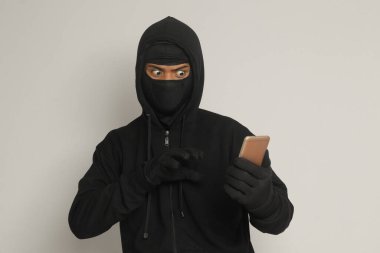 Siyah kapüşonlu ve maskeli gizemli bir adamın portresi cep telefonunda hackleme faaliyetleri yapıyor, elinde akıllı telefon tutan bir bilgisayar korsanı. Gri arkaplanda izole edilmiş resim