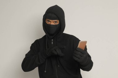 Siyah kapüşonlu ve maskeli gizemli bir adamın portresi cep telefonunda hackleme faaliyetleri yapıyor, elinde akıllı telefon tutan bir bilgisayar korsanı. Gri arkaplanda izole edilmiş resim