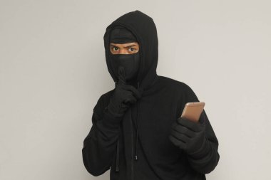 Siyah kapüşonlu ve maskeli gizemli bir adamın portresi cep telefonunda hackleme faaliyetleri yapıyor, elinde akıllı telefon tutan bir bilgisayar korsanı. Gri arkaplanda izole edilmiş resim