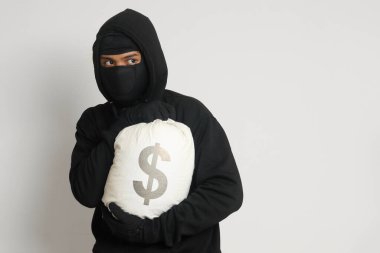 Siyah kapüşonlu ve maskeli gizemli hırsız para dolu bir çanta taşıyor. Gri arkaplanda izole edilmiş resim