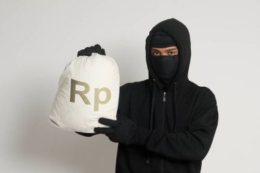 Siyah kapüşonlu ve maskeli gizemli hırsız bir çanta dolusu rupiah parası taşıyor. Gri arkaplanda izole edilmiş resim