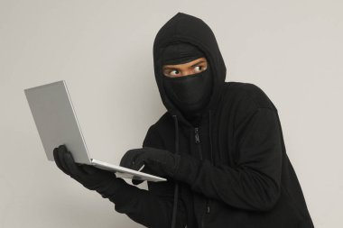 Siyah kapüşonlu ve maskeli gizemli bir adamın portresi. Bilgisayarda hackleme faaliyetleri yapıyor. Kişisel bilgisayar tutan bir bilgisayar korsanı. Siber güvenlik kavramı. Gri arkaplanda izole edilmiş resim