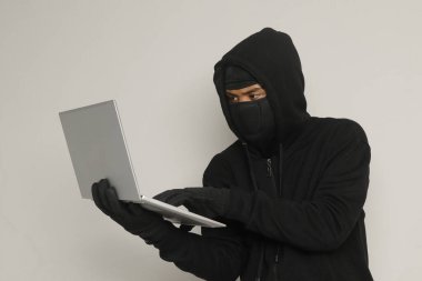 Siyah kapüşonlu ve maskeli gizemli bir adamın portresi. Bilgisayarda hackleme faaliyetleri yapıyor. Kişisel bilgisayar tutan bir bilgisayar korsanı. Siber güvenlik kavramı. Gri arkaplanda izole edilmiş resim