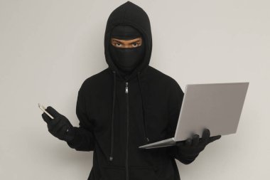 Siyah kapüşonlu ve maskeli gizemli bir adamın portresi. Bilgisayarda hackleme faaliyetleri yapıyor. Kişisel bilgisayar tutan bir bilgisayar korsanı. Siber güvenlik kavramı. Gri arkaplanda izole edilmiş resim