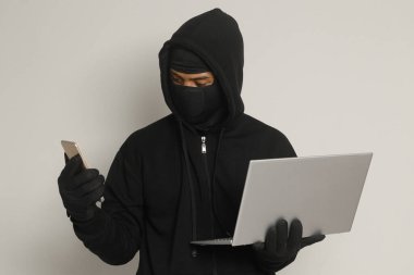 Siyah kapüşonlu ve maskeli gizemli bir adamın portresi dizüstü bilgisayar korsanlığı yapıyor, elinde cep telefonu olan bir bilgisayar korsanı. Siber güvenlik kavramı. Gri arkaplanda izole edilmiş resim