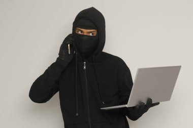 Siyah kapüşonlu ve maskeli gizemli bir adamın portresi dizüstü bilgisayar korsanlığı yapıyor, elinde cep telefonu olan bir bilgisayar korsanı. Siber güvenlik kavramı. Gri arkaplanda izole edilmiş resim