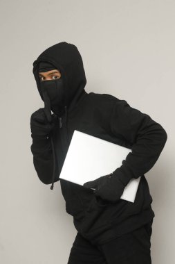 Siyah kapüşonlu ve maskeli gizemli hırsız dizüstü bilgisayar çalıp sıvışıyor. Gri arkaplanda izole edilmiş resim