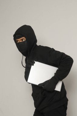 Siyah kapüşonlu ve maskeli gizemli hırsız dizüstü bilgisayar çalıp sıvışıyor. Gri arkaplanda izole edilmiş resim