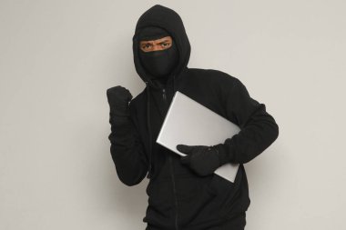 Siyah kapüşonlu ve maskeli gizemli hırsız yumruğunu kaldırıp başarıyı kutlarken dizüstü bilgisayar çalıyor. Gri arkaplanda izole edilmiş resim